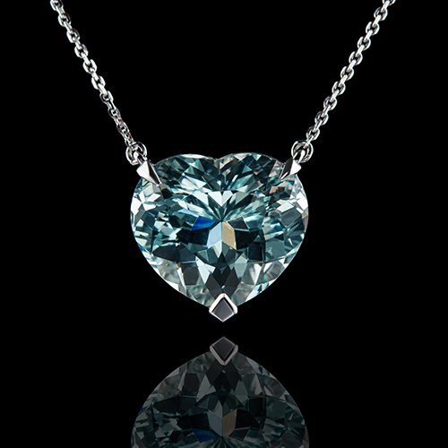 Heart-shaped light blue aquamarine gemstone pendant on a silver chain displayed on a reflective black surface.