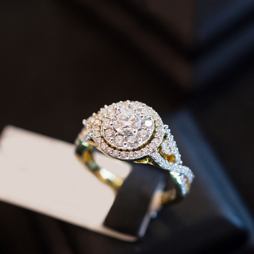 Close-up of a two-tone gold round diamond engagement ring featuring a double halo and a twisted pav&eacute; band on a display stand.