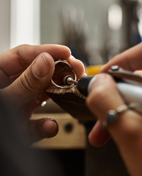 Close-up of a master jeweler's hands using a rotary polishing tool to finish a custom ring on a wooden jeweler's bench pin.