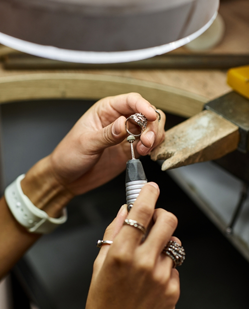 Close-up of a professional bench jeweler using a precision rotary tool to craft a textured silver ring under a bright workbench lamp.